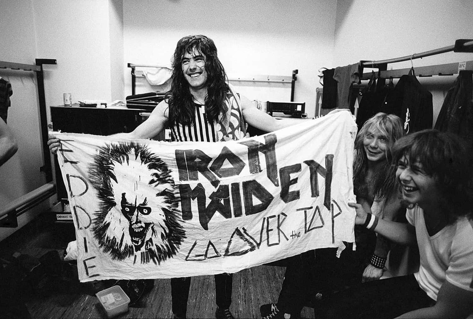 Iron Maiden backstage in 1980, Paul Di'Anno holding up a hand-painted fan banner reading Iron Maiden Go Over the Top with an early Eddie drawing, band members laughing behind him.