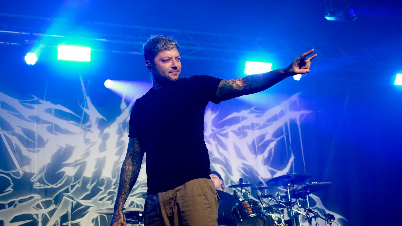 Chelsea Grin's vocalist points out into the crowd from the front of the stage under blue and green lights, the band's logo backdrop and drum kit behind him, New Century Hall, Manchester, December 2025.