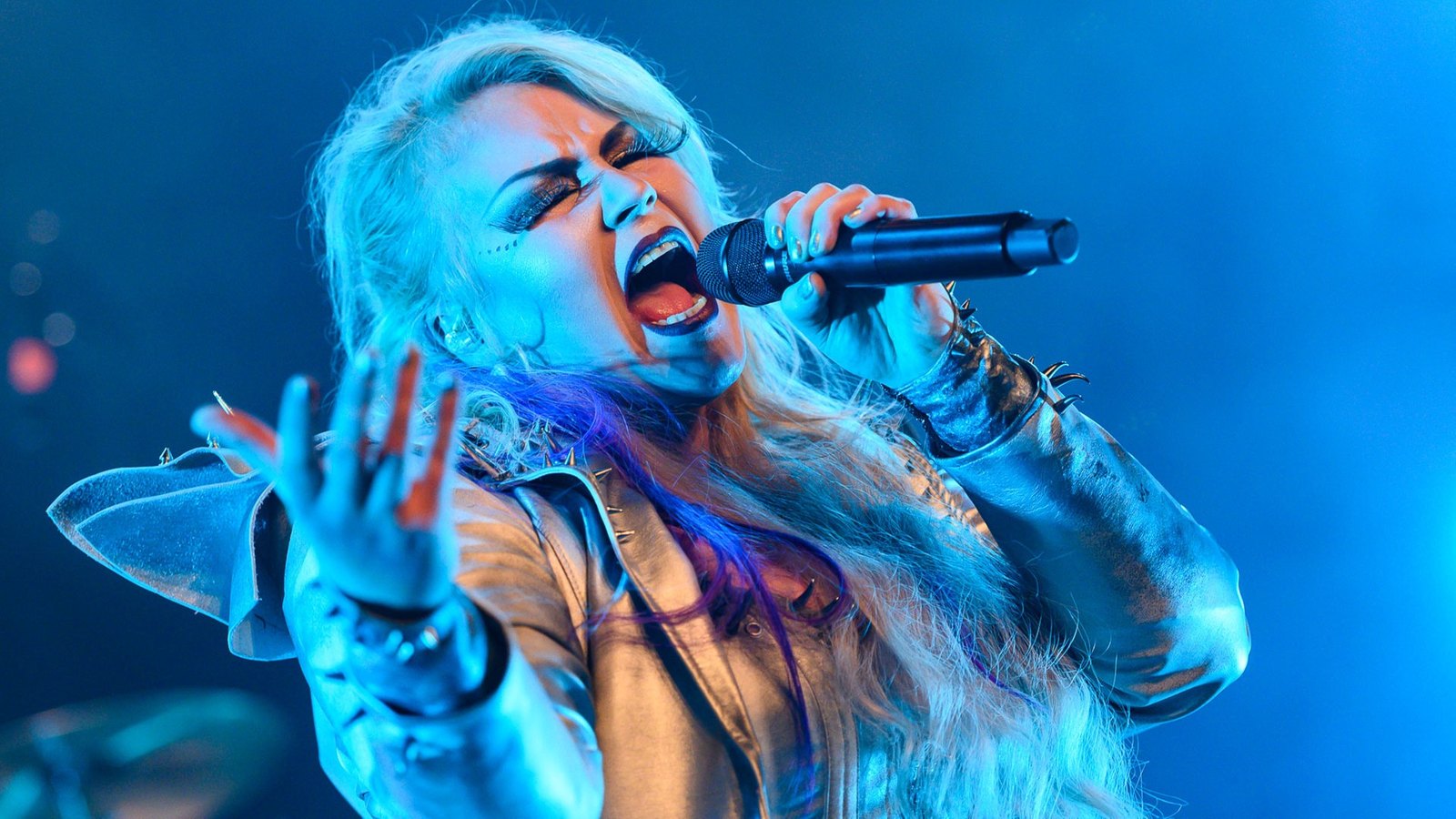 A Battle Beast singer with eyes shut and mouth wide open, caught mid-phrase with a microphone in one hand and the other hand stretched out, in blue stage light