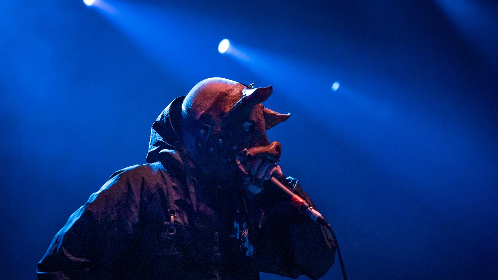 To the Grave's vocalist in a pig mask and black hooded jacket, hunched over the microphone mid-vocal under blue stage light.