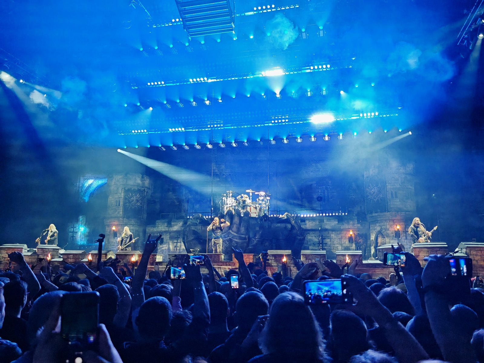 Sabaton on the stage of Tauron Arena in Kraków in 2025, the band small in front of a castle set under rigs of lights, the crowd's heads and raised phones filling the foreground
