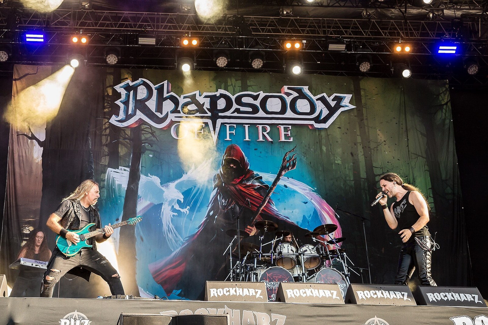 Rhapsody of Fire at Rockharz Open Air 2025: a guitarist leans back over his instrument at the left, the drummer plays at the centre under the band's painted backdrop, and the singer holds the microphone at the right.