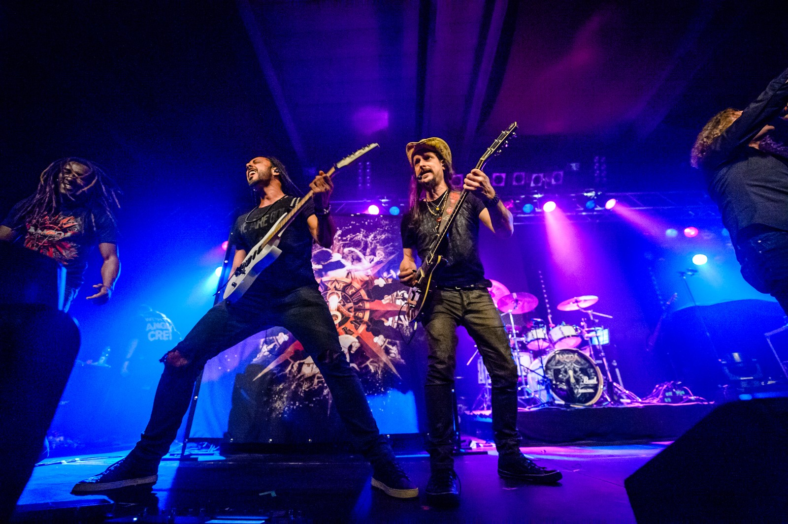 Two guitarists of Angra side by side on stage at the Live Music Hall in Cologne in 2016, with the drum kit and the band's backdrop behind them