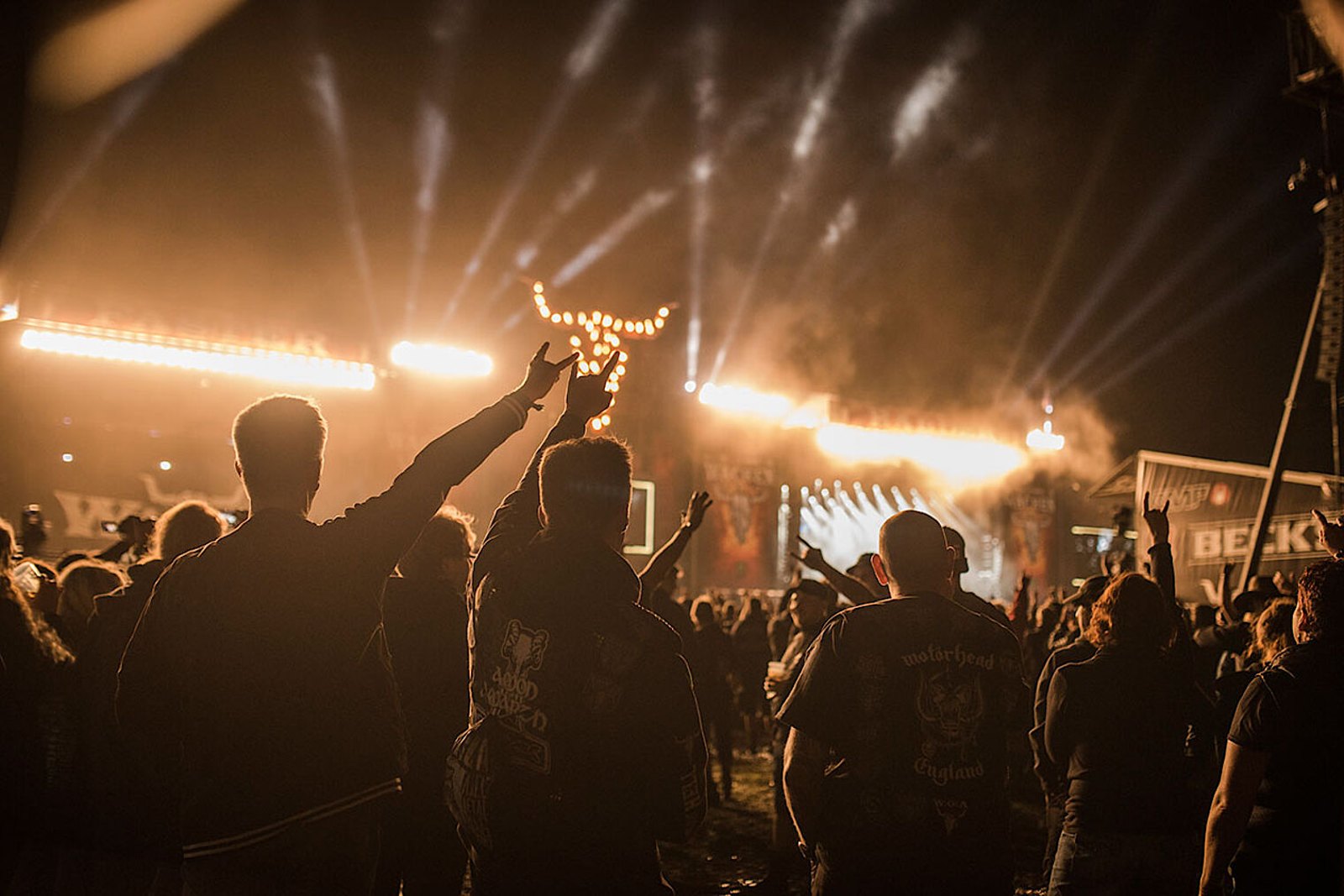 A festival crowd seen from behind, fists and horns raised, the lit stage and pyrotechnics beyond them