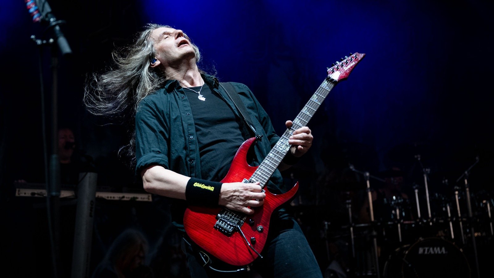 A Blind Guardian guitarist with his head thrown back, playing a red Ibanez on the Rock Hard Festival stage in Gelsenkirchen in 2022, a Blind Guardian wristband on his picking arm
