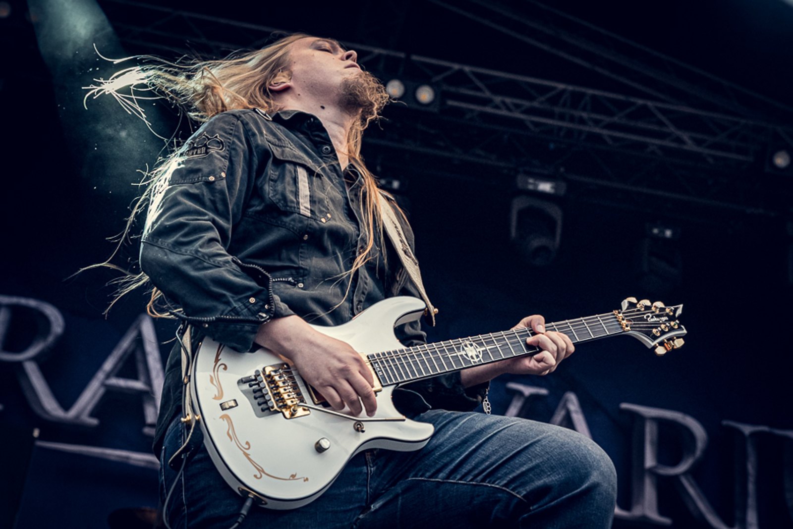 A Stratovarius guitarist playing a white guitar on stage, the band's name on the backdrop behind him