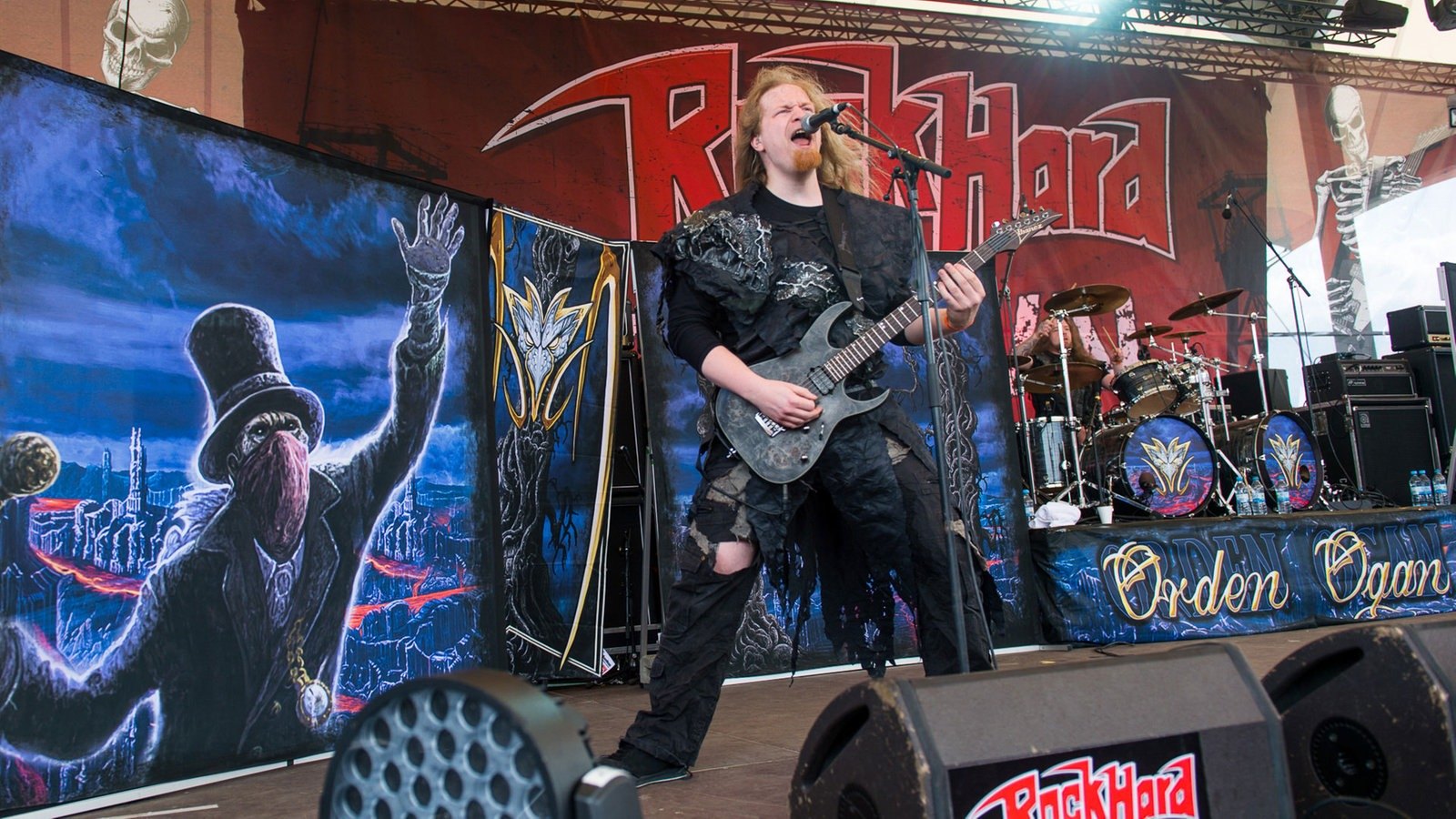 An Orden Ogan guitarist singing at the microphone on the Rock Hard Festival stage in 2016, the band's painted banners and a drum riser lettered Orden Ogan behind him