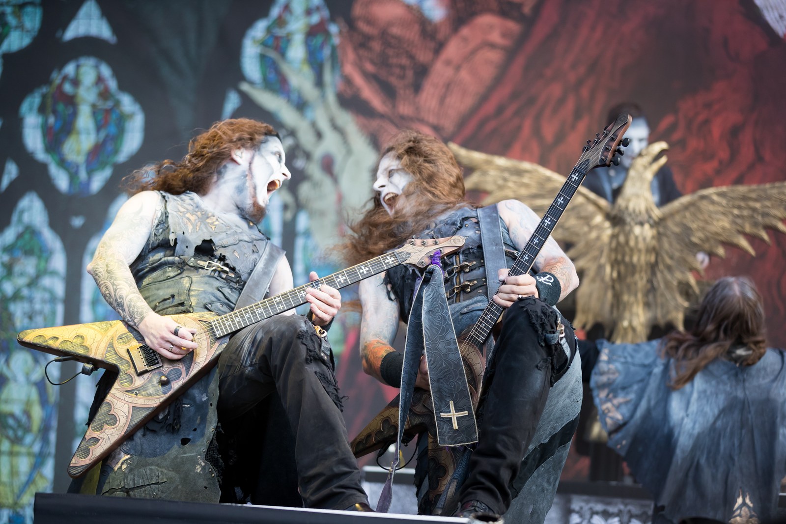 Two Powerwolf guitarists face to face on stage at Wacken Open Air in 2017, both roaring as they play, with the band's stained-glass backdrop and a golden eagle behind them