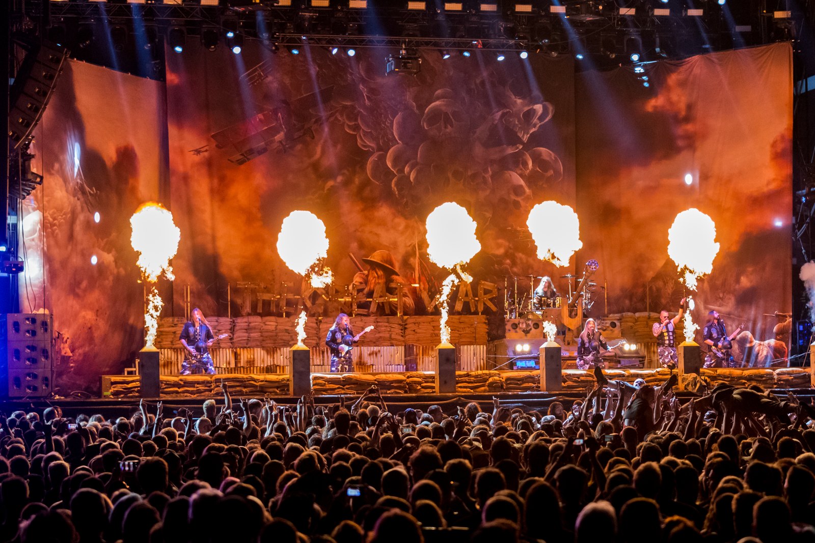 Sabaton on the Wacken Open Air stage at night, flame jets firing along a sandbagged stage set under a painted backdrop of skulls, with the crowd's raised hands filling the foreground