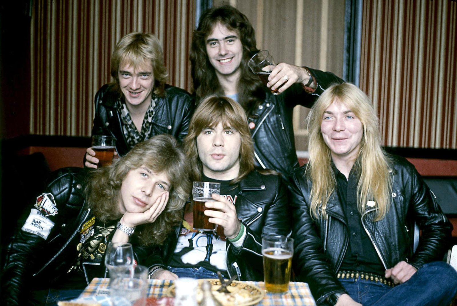 Iron Maiden's five members, the Paul Di'Anno-era line-up with Steve Harris and Dave Murray, crowded around a pub table, laughing and holding pints, an Iron Maiden backstage laminate on one jacket