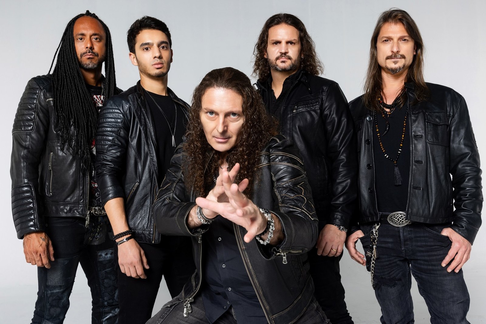 The five members of Angra standing together in leather jackets for a studio group portrait, the singer crouched at the front with one hand held out
