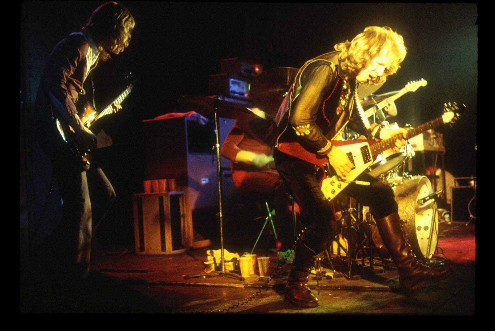 Two Wishbone Ash guitarists playing at opposite sides of a small stage, the drummer between them behind the kit