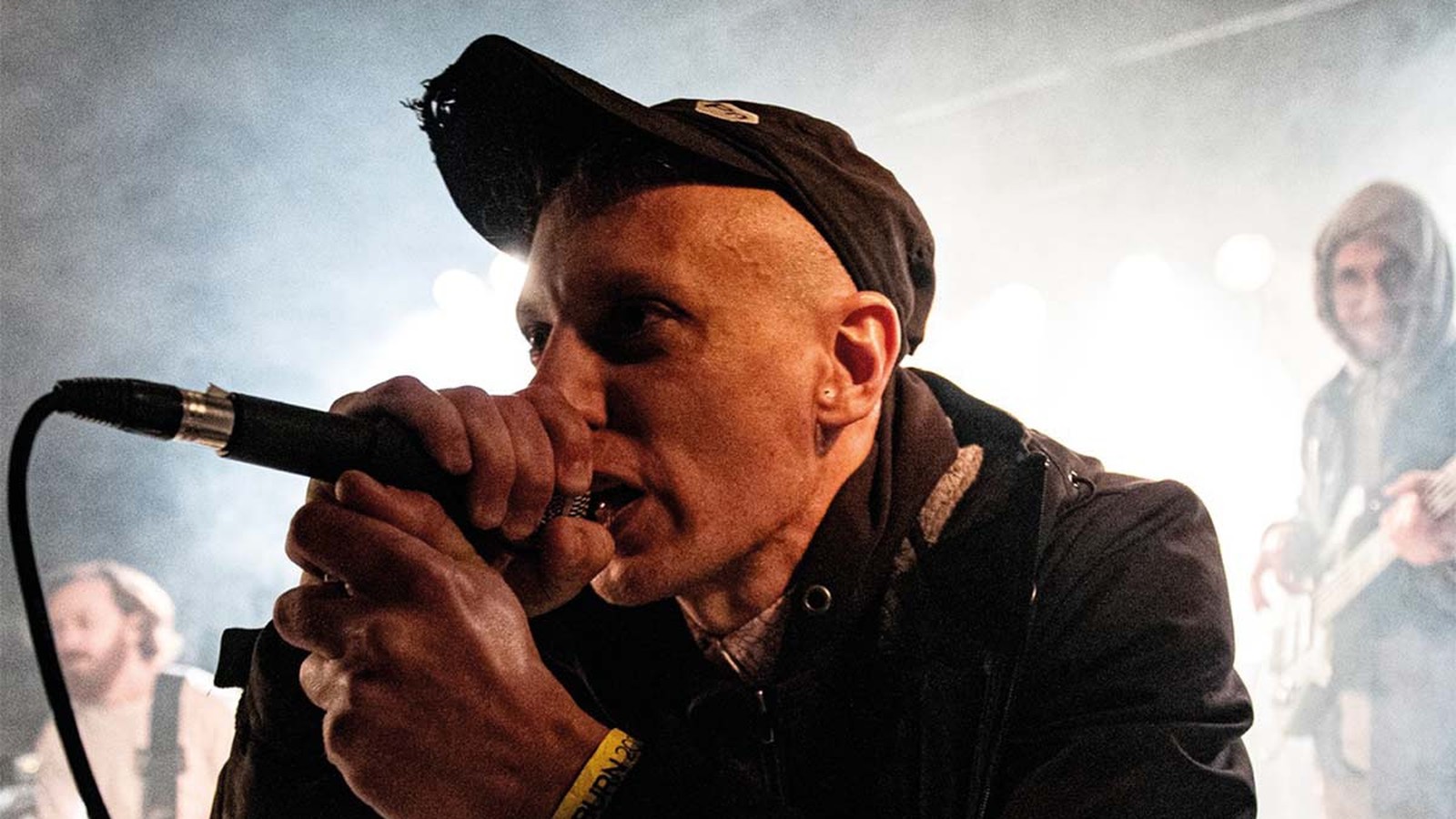 Thou singer Bryan Funck in a cap gripping the microphone with both hands in stage smoke, a guitarist behind him