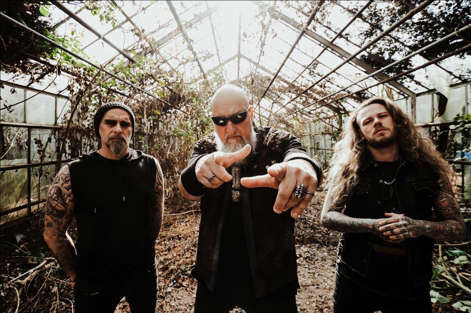 The three members of Rage standing in a derelict greenhouse, Peavy Wagner in the middle with both hands raised towards the camera