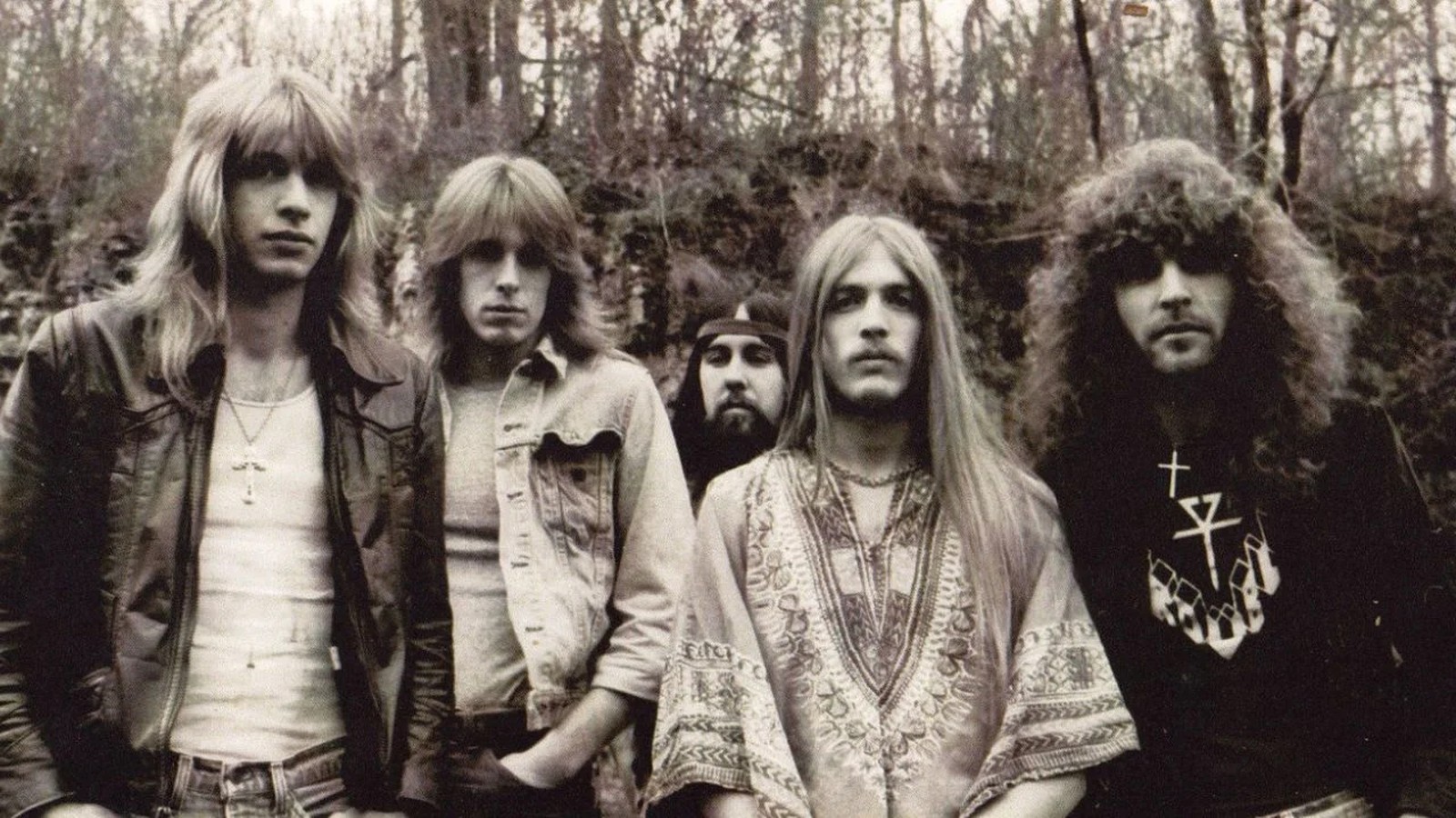 The five members of Trouble with long hair and denim and leather jackets, posed in front of trees in a vintage black and white photograph