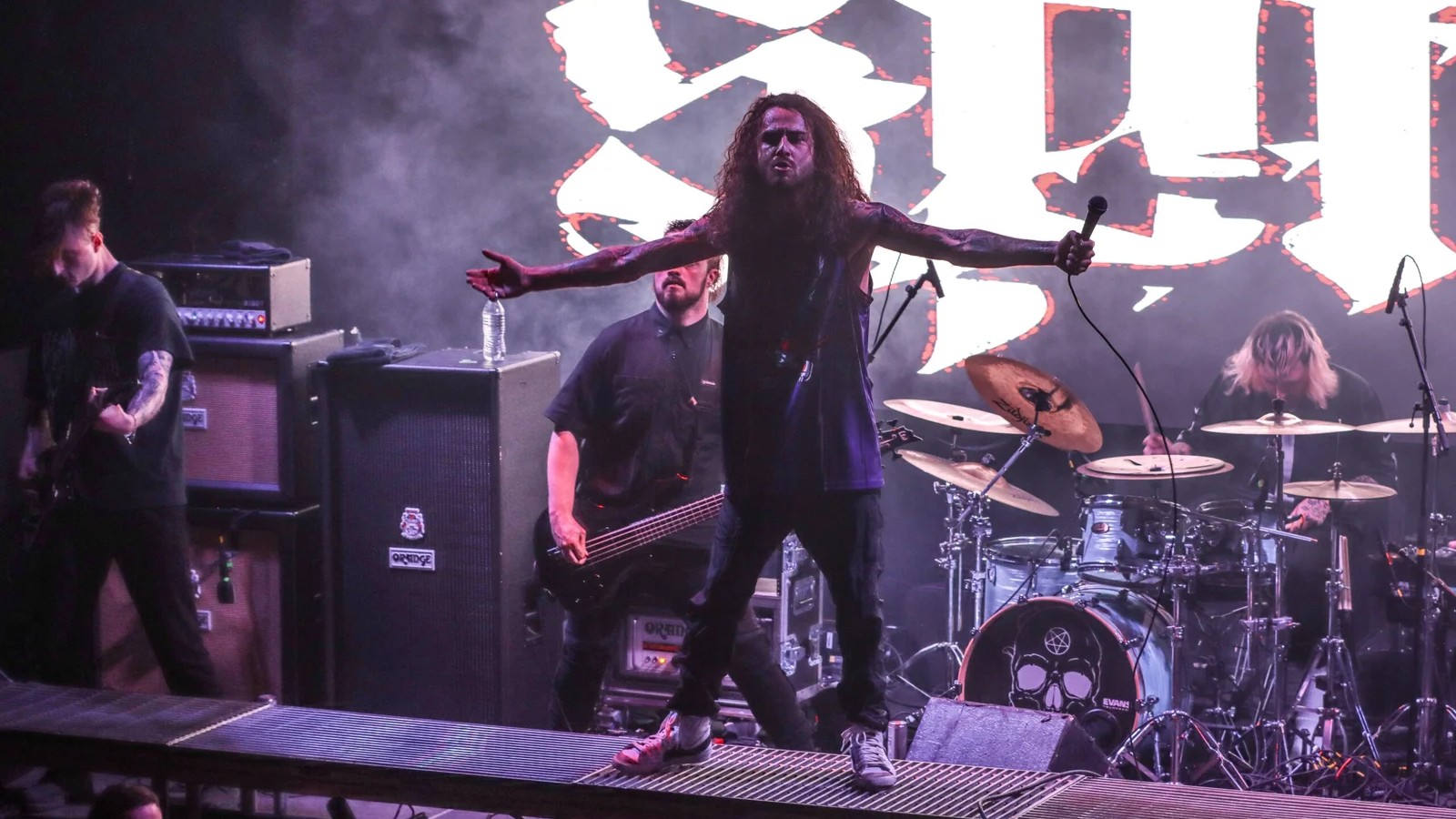 Spite vocalist standing arms outstretched on a stage riser in front of the band's logo on a video backdrop, the band playing behind him in purple light and smoke.