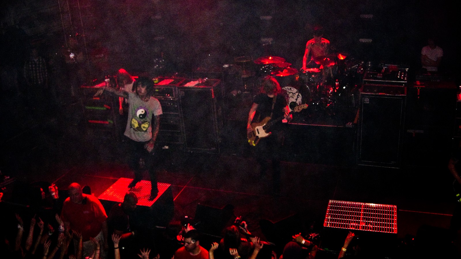 Bring Me the Horizon on a dark, smoke-filled stage in red light, the vocalist on a riser with an arm raised toward a crowd of raised hands, the bassist beside him and the drummer behind