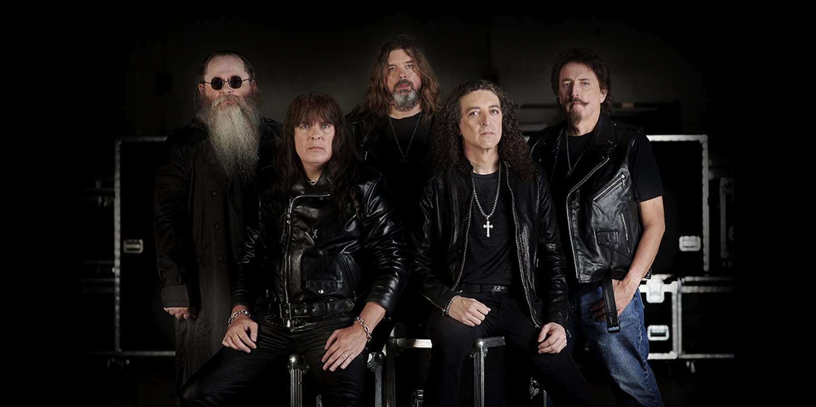 The five members of Jag Panzer in black leather, seated and standing in front of stacked flight cases for a studio portrait