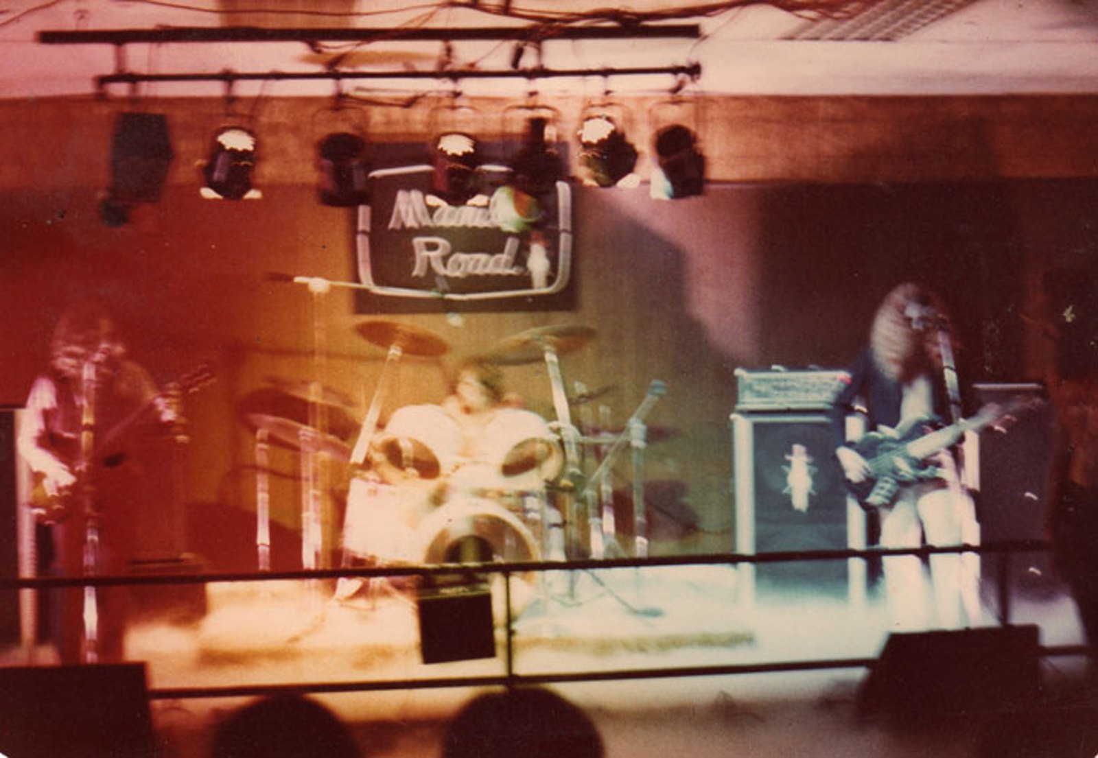 Manilla Road playing a small club stage, a lit sign with the band name hanging above the drum kit, guitarist and bassist either side of it