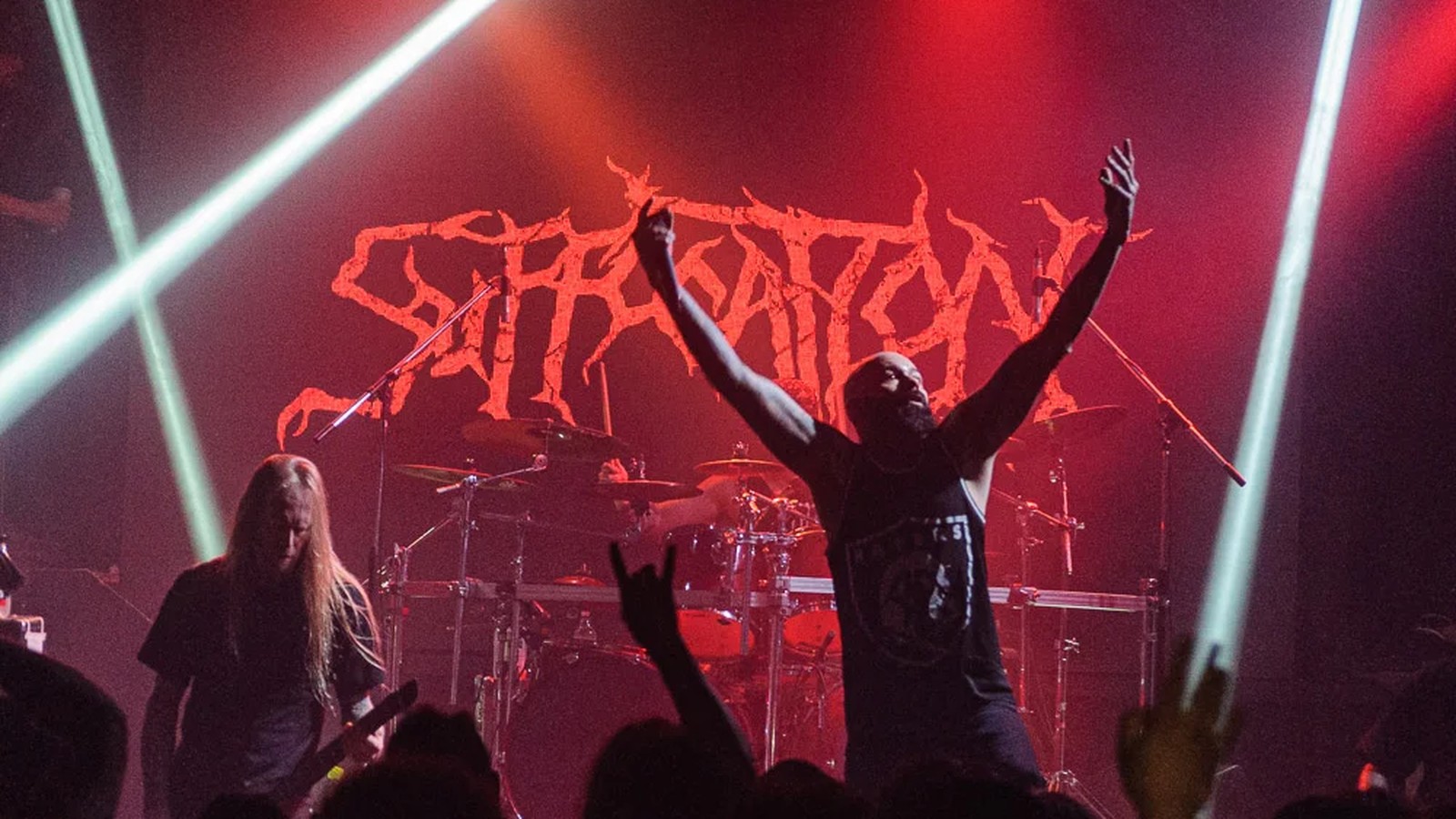 Suffocation vocalist with both arms raised before the band's red logo backdrop and drum kit, a guitarist at left and crowd silhouettes in front, live at El Teatrito in Buenos Aires.