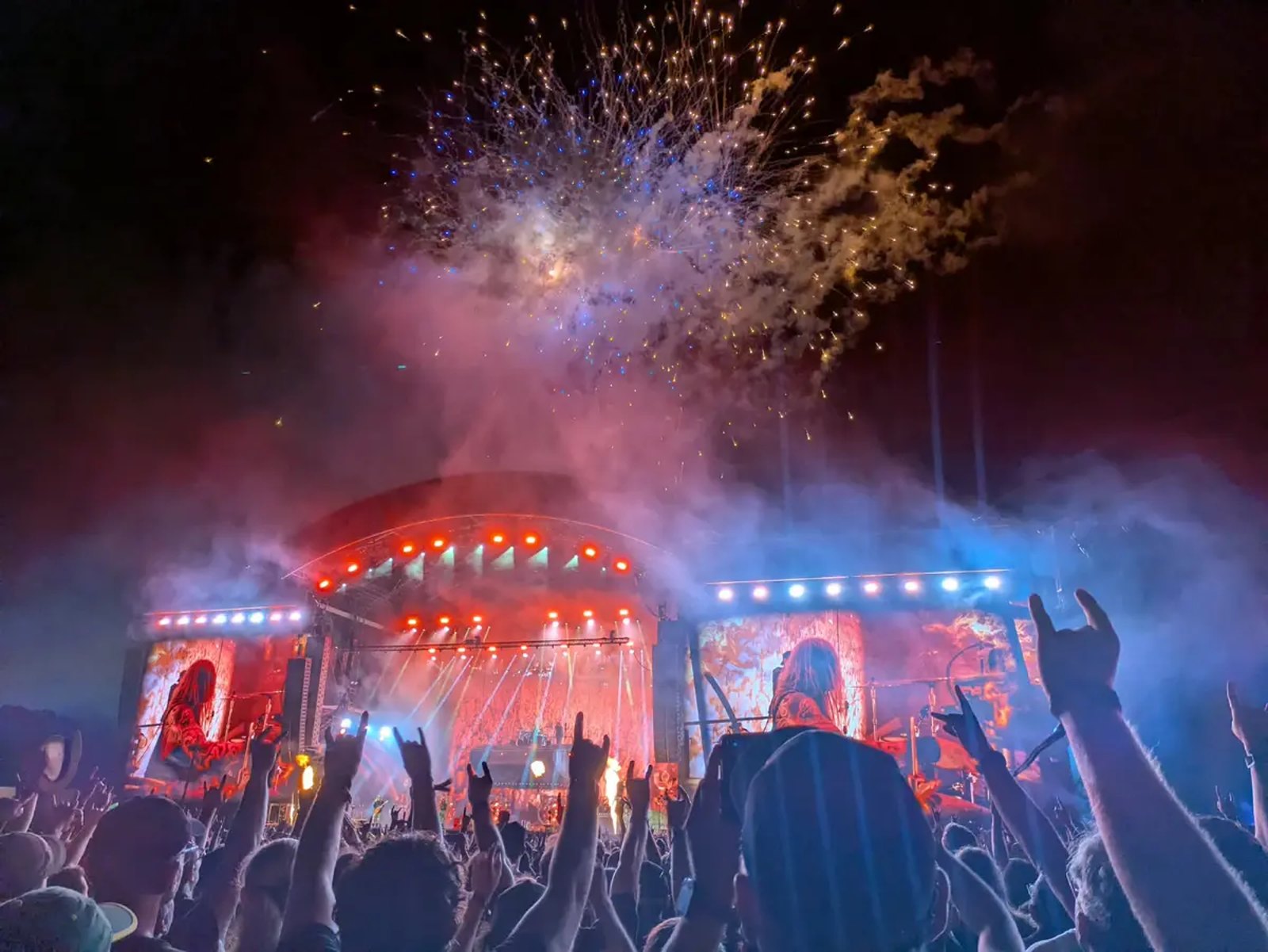 A festival crowd with arms and horns raised in the foreground, the lit stage beyond them and fireworks bursting overhead